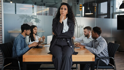 Pensive thinking thoughtful businesswoman female leader boss woman sitting on table in boardroom think about project idea on background of multicultural employees workers colleagues work meeting