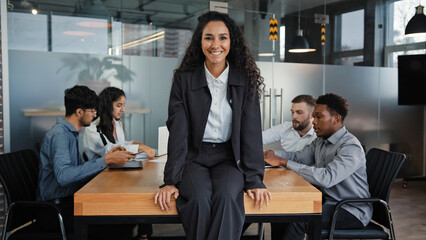 Satisfied smiling female leader boss woman company CEO businesswoman sitting on table in office smile to camera background of multiracial coworkers colleagues teamwork project startup analyze work