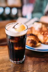 Ice coffee in a tall glass with cream poured over on a old rustic wooden table with blurred pastries on background. Cold summer drink with copy space.