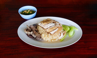 Boiled chicken, offal with cucumber sliced on white dish and small bowl spicy or sweet sauce on dark brown wooden background. Steamed rice topped with chicken and fresh vegetable on table. Asian food.