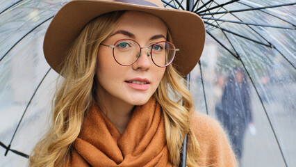 Close-up Caucasian 20s girl in eyeglasses and hat posing on street in city with transparent...