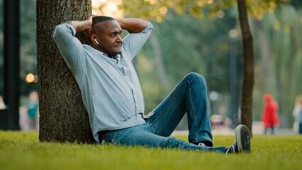 Happy calm relaxed elderly middle-aged mature senior adult African American ethnic man in wireless headphones sitting on grass in city park under tree listen music audio song in smartphone outdoors