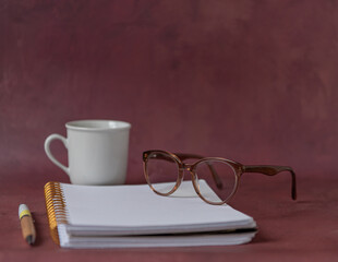 Cahier ouvert  et tasse blanche sur une page blanche, lunettes et stylo sur un gond rouge
