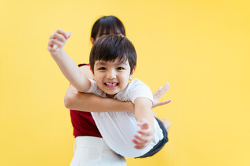 Happy cheerful Asian woman and a little young boy playing together, a woman piggyback or carrying a little boy on her back. Woman and boy portrait on yellow pastel background.