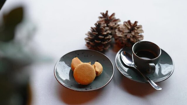 Golden macarons served with a cup of hot coffee on white table decorated with pine cones. Festive desserts. Christmas baking. Horizontal indoor shot. High quality 4k footage