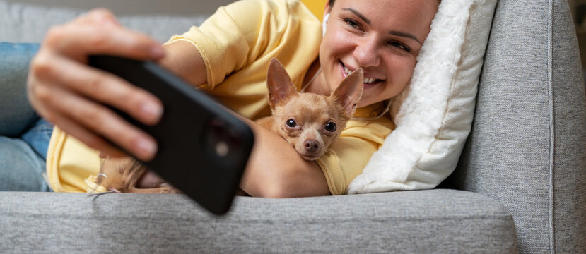 Young brunette woman taking selfie using her smartphone while lying down on sofa with little dog.