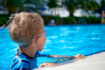 Child is swimming in a clean outdoor pool, the child's head above the water, turned away from the camera. Boy swims independently in the blue transparent water of the summer pool