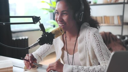 Young cheerful sociable African American woman makes notes in notebook and puts on headphones to talk with friends or followers from social networks dressed in casual style sits at table in apartment - Powered by Adobe