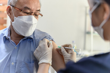 Close up hand of Asian woman nurse injecting covid-19 vaccine to senior Thai man patient wearing mask in clinic or health care center. Coronavirus pandemic protection or health care medical concept.