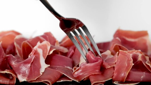Prosciutto crudo or jamon meat on serving board on white background. Hand with fork takes Prosciutto. Sliced into thin pieces of ham on cutting serving  board. Italian antipasto 