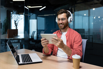 Successful businessman at workplace smiling man watching online video sitting at desk wearing headphones and glasses programmer holding tablet computer inside office.