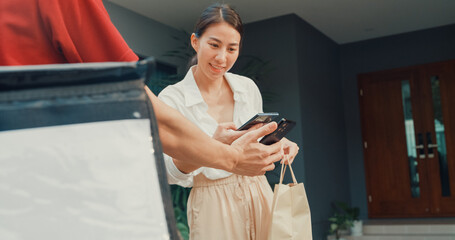 Young happy Asian woman accept paper bag from delivery man with bicycle in red uniform carry case box scan QR code payment on smartphone in front of house. express food delivery concept.