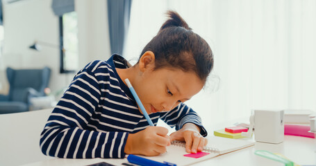 Asian toddler girl with sweater sit in front of desk with notepad use pencil focus on write notebook do homework from online learning course on the weekend at home. Distance online learning concept.