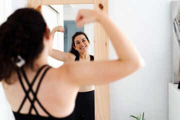 Funny young woman flexing arms looking in the mirror after training