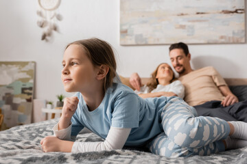 cheerful girl lying on bed near blurred parents resting on background.