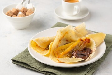 Thin pancakes stuffed with caramelized bananas and chocolate spread on a white plate against a light concrete background. International pancake day.