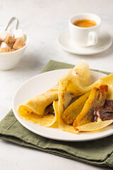 Thin pancakes stuffed with caramelized bananas and chocolate spread on a white plate against a light concrete background. International pancake day.