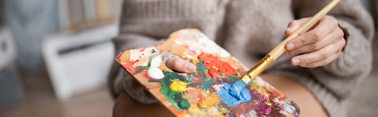 Cropped view of artist in sweater holding paintbrush and palette in workshop, banner