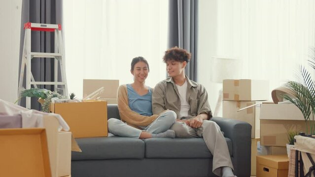 Young Happy Asian Sweet Couple Sit On Sofa Couch Smiling Looking At Camera Show Proud Show Off New House Keychain In Living Room. Husband Wife Move New Home, Apartment Rent And Ownership Concept.