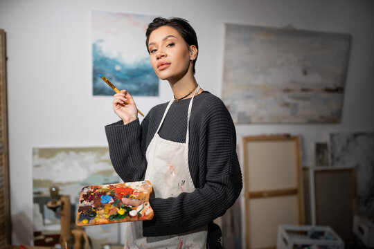 Brunette artist looking at camera while holding paintbrush and palette in blurred workshop