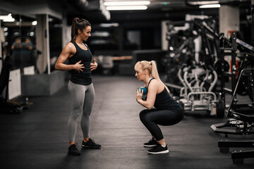 Fototapeta na wymiar A female personal trainer is explaining how to do exercises to a sportswoman who is squatting and holding kettle bell.
