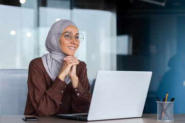 Smiling and dreamy businesswoman working inside office with laptop, woman in hijab and glasses...