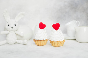 two cakes with red hearts, white knitted bunny and teapot, cups on a gray background.