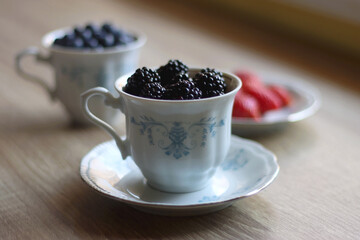 Vintage porcelain cups and plates filled with various fresh berries. Wooden background, selective focus.