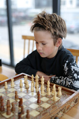 a boy of eight years, Caucasian appearance, sits in a bright room, plays chess with a friend. The boy is focused, thinks through the moves