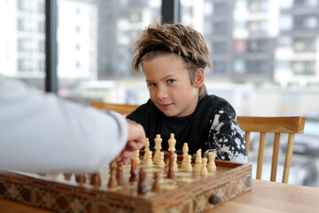 a boy of eight years, Caucasian appearance, sits in a bright room, plays chess with a friend. The boy is focused, thinks through the moves