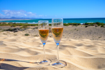 Two glasses of rose champagne or cava sparkling wine served on white sandy tropical beach with dunes and blue ocean, romantic vacation, winter sun on Fuerteventura, Canary, Spain