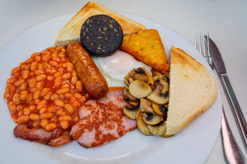 White board with full English breakfast with bacon, fried egg, beans, tomato, roasted sausage, black pudding, scons, hash browns and fried mushrooms