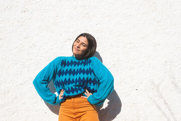 Smiling young latin girl in colorful clothes, saturated blue argyle sweater and orange pants, white stucco wall background outside, light sunny day. Copy space on one side