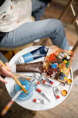 Top view of artist in apron putting paintbrush in jar with water near paints on table in workshop