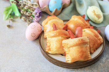 Buns in the form of an Easter rabbit from sausage and cheese in a yeast dough with colored eggs on a gray stone background. Traditional Easter symbols and food. Copy space.