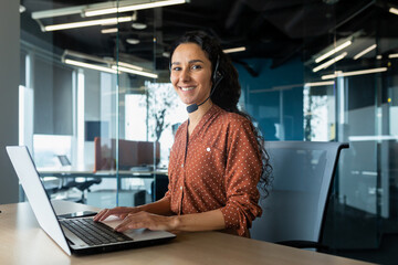 Portrait of Latin American business woman, office worker looking at camera and smiling, using headset and laptop for remote online communication, customer support tech call center worker. - obrazy, fototapety, plakaty