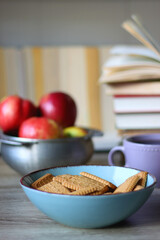 Stack of books, e-reader, reading glasses, bowl of biscuits, apples and cup of tea on the table. Bookshelf in the background. Selective focus.