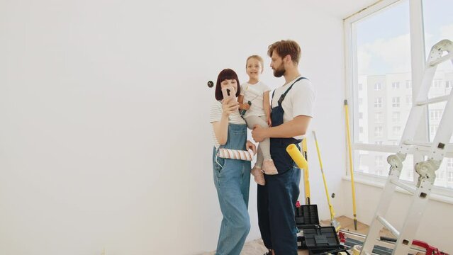 A cheerful happy family is renovating a purchased apartment. A very smiling family of three people taking a selfie. The child hugs his parents tightly. In the background, repairs are taking place