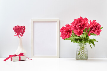 Mockup with a white frame and red peonies in a vase, red heart and gift box on a white table. Empty poster frame mockup for presentation, design, lettering. Valentines Day, Happy Women's Day concept