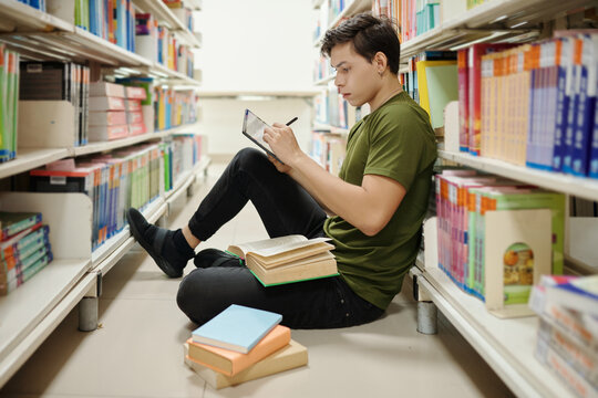Smart college student sitting on library floor and writing essay on tablet computer