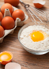 Bowl plate with flour and egg yolk on wooden background with fresh raw eggs and metal whisk