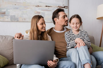 happy woman sitting with laptop near husband kissing daughter in living room.
