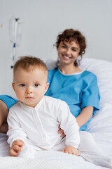 toddler girl in romper looking at camera near blurred mother smiling on hospital bed.