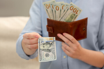 Woman giving 100 dollar bill and holding brown leather wallet with banknotes indoors, closeup. Money exchange