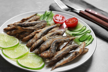 Plate with delicious fried anchovies, lime, cucumber and tomato on grey table, closeup