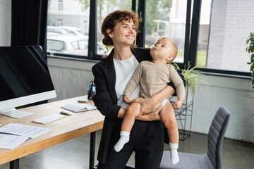 happy businesswoman looking away while standing with little child near computer monitor with blank screen.