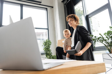 happy businesswoman in black suit holding toddler child and notebook near laptop on blurred foreground.
