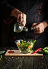 Professional chef adds olive oil on a salad of fresh vegetables in a plate on a wooden board. Cooking healthy food in the kitchen with the hands of a cook