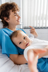 happy woman smiling on hospital bed near baby looking at camera on blurred foreground.