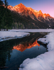 Snowcapped Alps mountains reflected with warm colors in the partially frozen Anterselva lake (North Italy) at sunset in winter. Snow and ice in the foreground, tree and forest in the background.